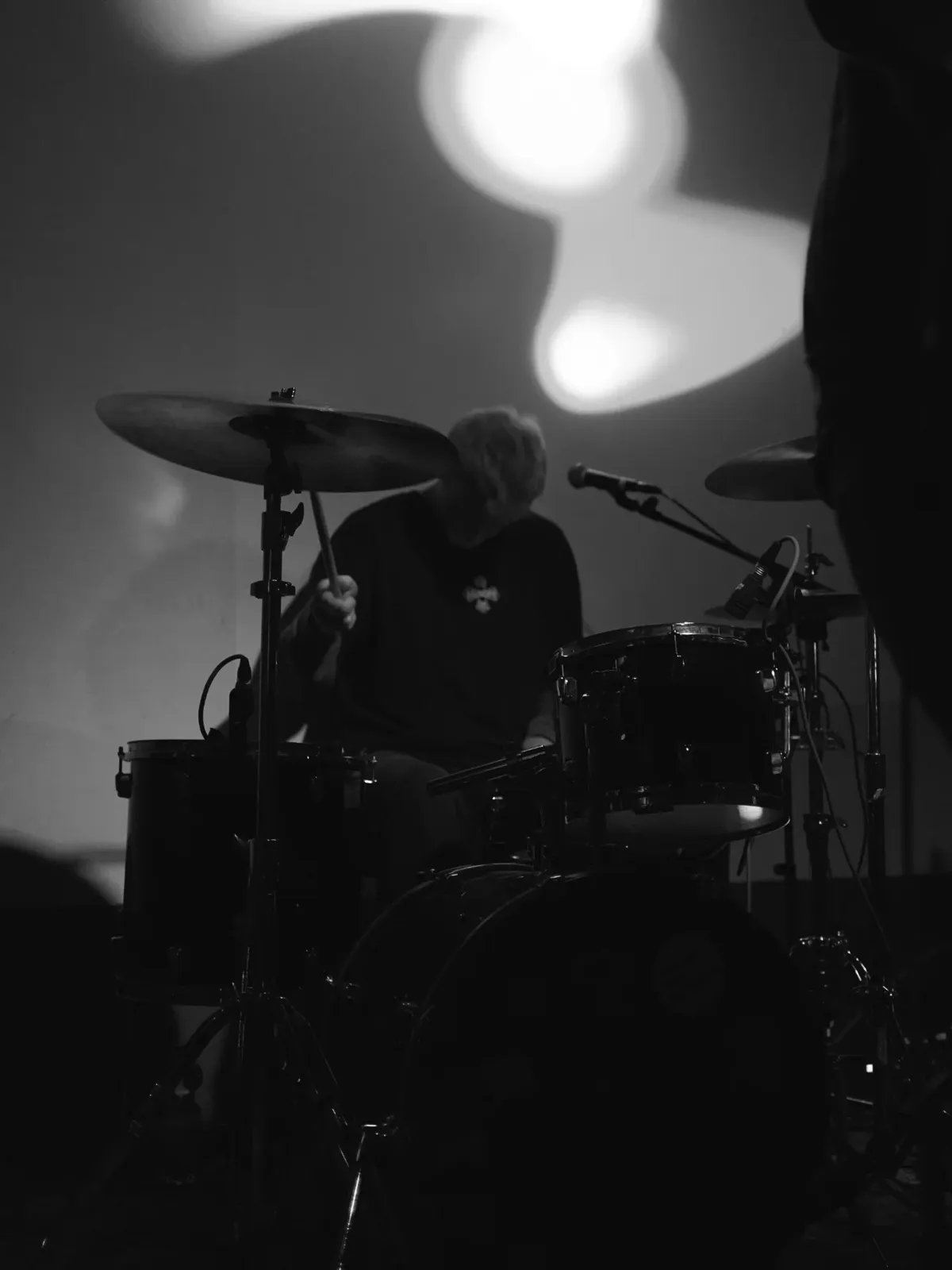 Black and white: the drummer behind the kit, half in shadow, a blur of light on the wall behind