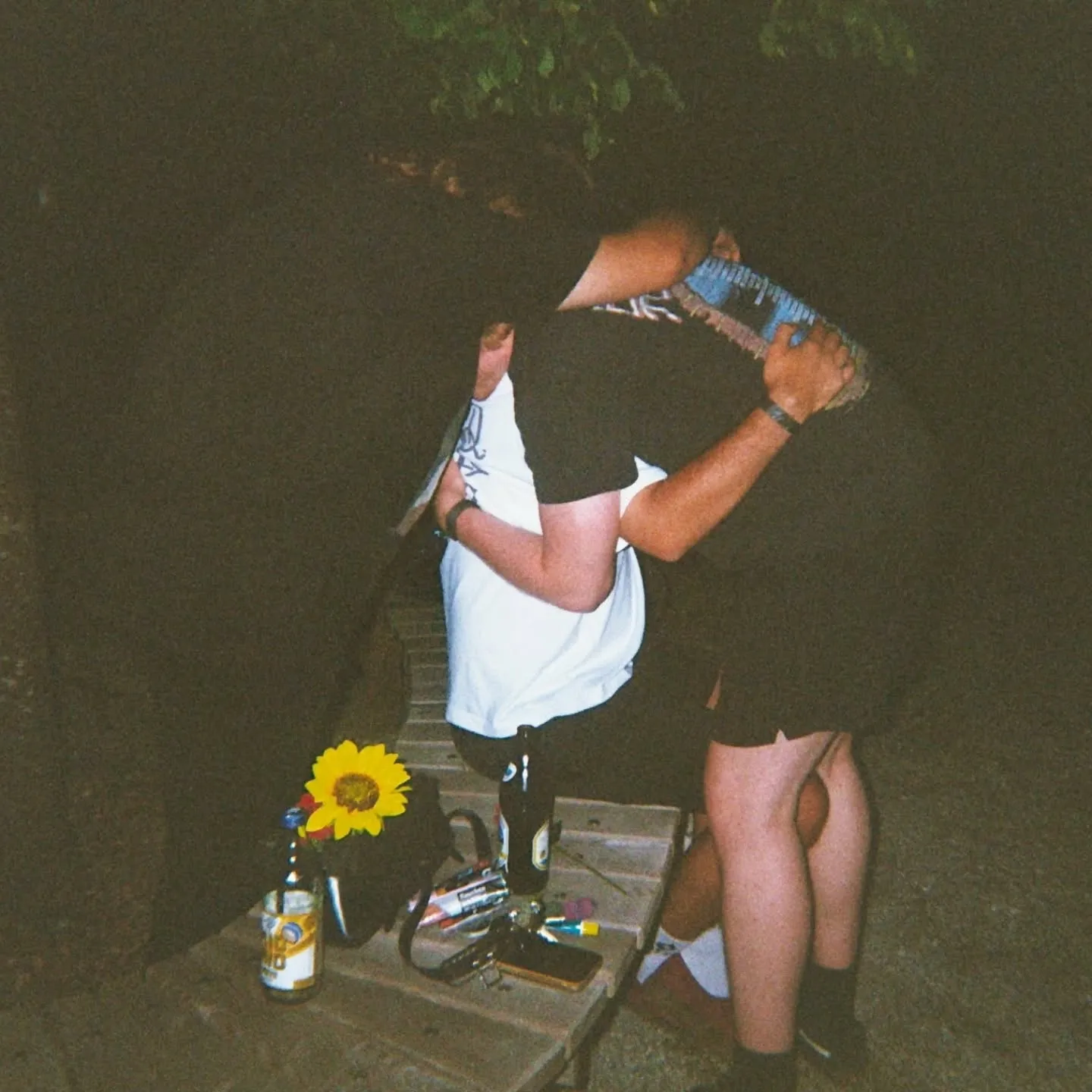 A flash photo outside at night: two people hugging by a wooden bench, a sunflower and beer bottles on the bench