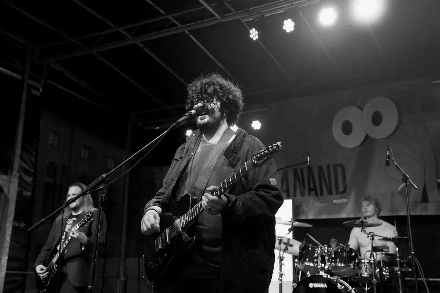 Black and white: singing at the microphone, guitar in hand, the drummer and a guitarist behind